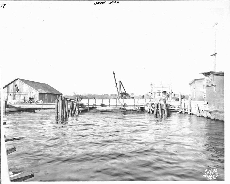 View of the west elevation of the concrete bridge during construction. At left, workers place wire mesh around the steel beams before they are encased in concrete. The piles at either side of the bascule leaf are placed to protect the span from impact. Sanborn Fire Insurance Maps identify the building at left as a fertilizer warehouse. Photo from the Maryland Department of Transportation.